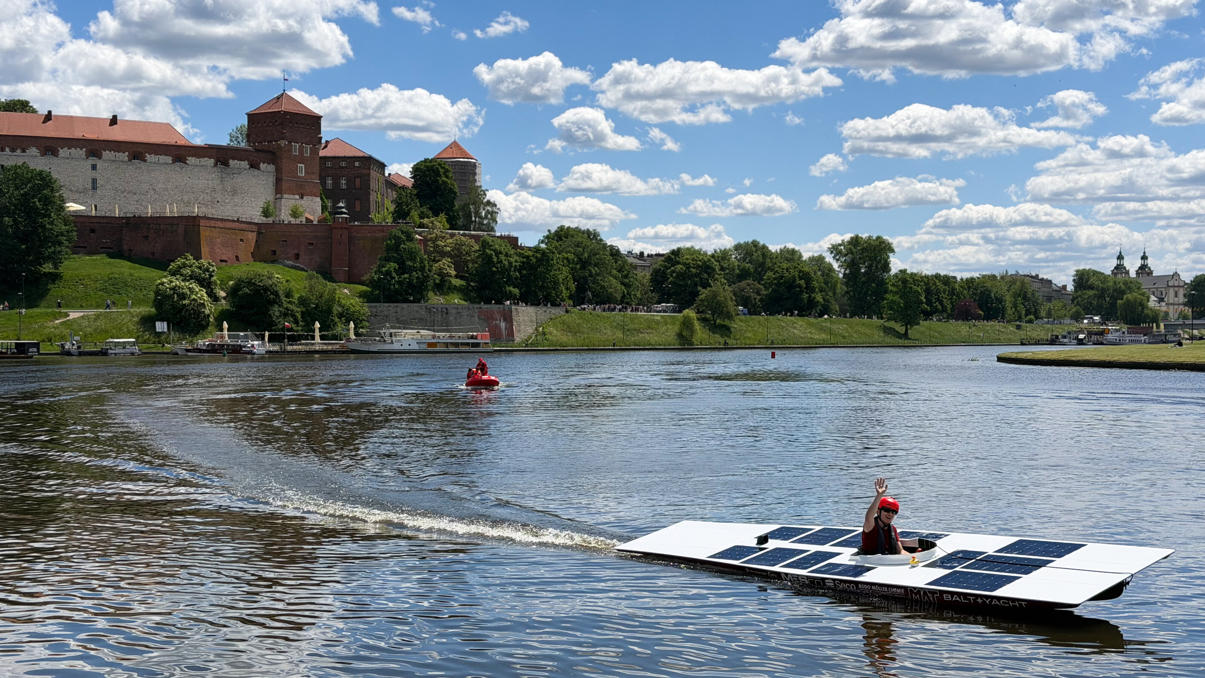Solarna Delta płynie po Wiśle w pobliżu Wawelu. Na pokładzie jedna osoba w kasku unosi w górę rękę.