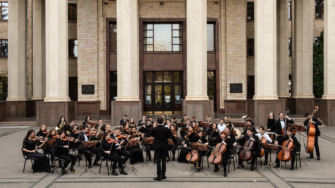 Orkiestra MASO złożona z ponad 40 instrumentalistów koncertująca na placu przed budynkiem. Większość instrumentów stanową skrzypce, flety i wiolonczele, z prawej strony dwa kontrabasy. Przed orkiestrą stoi dyrygent. 