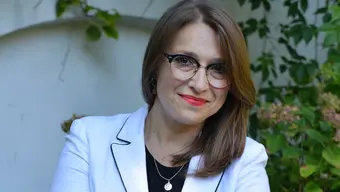 A portrait photo of the laureate, Marta Foryś. A smiling woman with long light brown hair with glasses. She wears a black blouse with a white jacket and a necklace. Behind her is a fragment of a wall and green plants.
