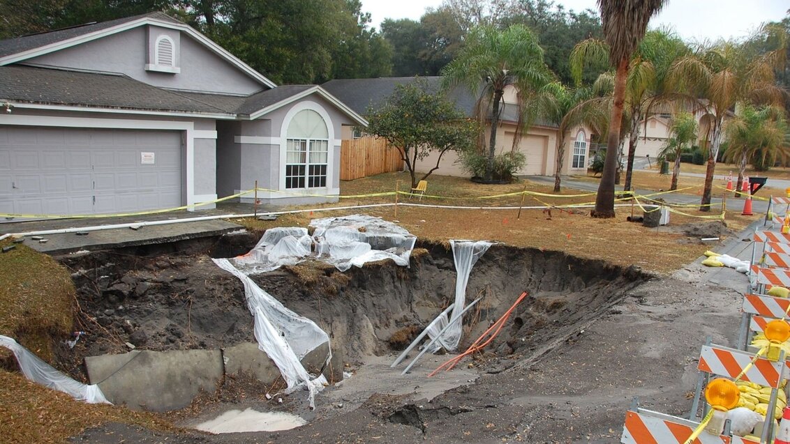 In the foreground, there is a large sinkhole formed in the front yard of a house. In the background, there is a single-storey greyish house with a sloped roof.