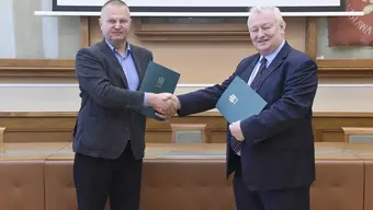 Na zdjęciu dwóch mężczyzn. Podają sobie prawą dłoń w uścisku. W lewej każdy z nich trzyma obwolutę na dokumenty.