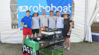 Six students standing next to each other, members of AGH Solar Boat. They wear t-shirts and shorts. In front of them, there is the “Barka” autonomous boat and behind them, a roll-up with the name of the competition.