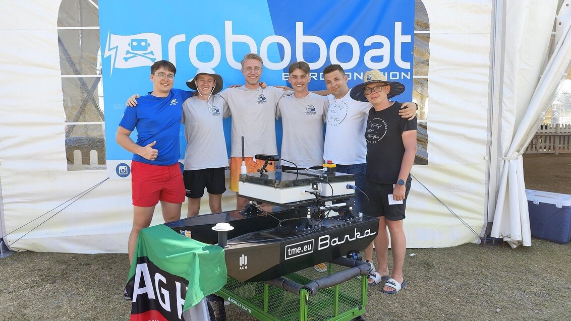 Na zdjęciu sześciu studentów - członków AGH Solar Boat. Ubrani w t-shirty i shorty, stoją obok siebie, obejmując się ramionami. Przed nimi autonomiczna łódź "Barka", za nimi ścianka z nazwą zawodów RoboBoat.