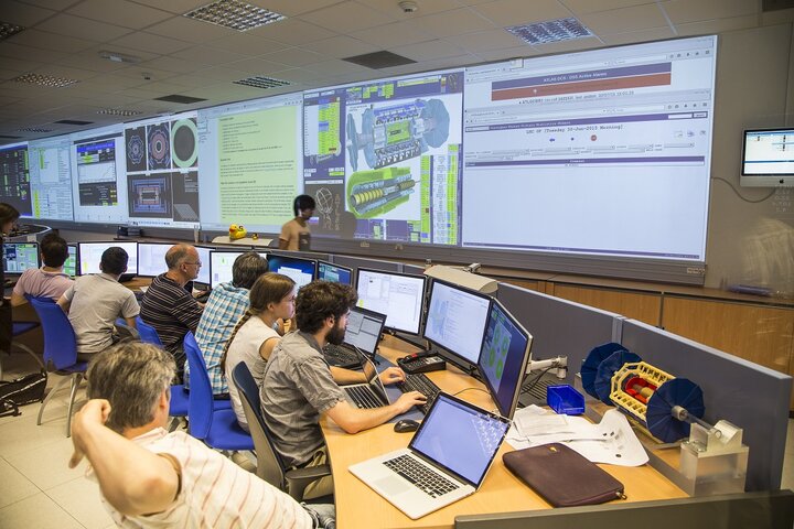 Zdjęcie przedstawia grupę naukowców siedzących przy komputerach. Na pierwszym planie widocznych jest sześciu mężczyzn i jedna kobieta. Na monitorach wyświetlają się różnego rodzaju wykresy. Na drugim planie, na ścianie wyświetlają się w dużym formacie (pięć pod rząd) slajdy z różnymi analizami.