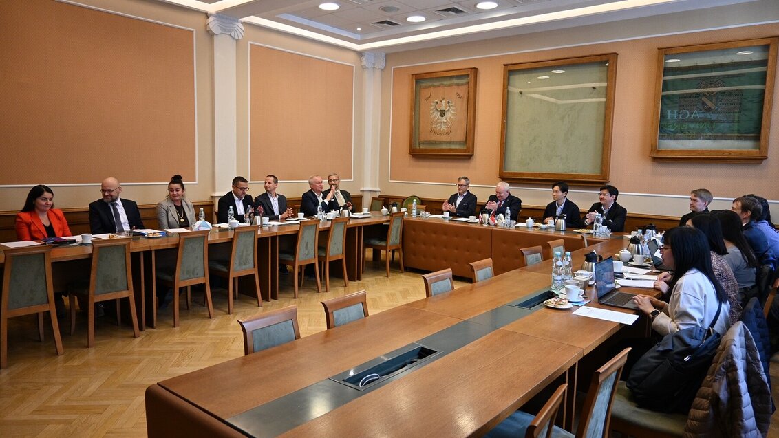 The image shows a meeting or conference room with a large group of people seated around a long, U-shaped table.