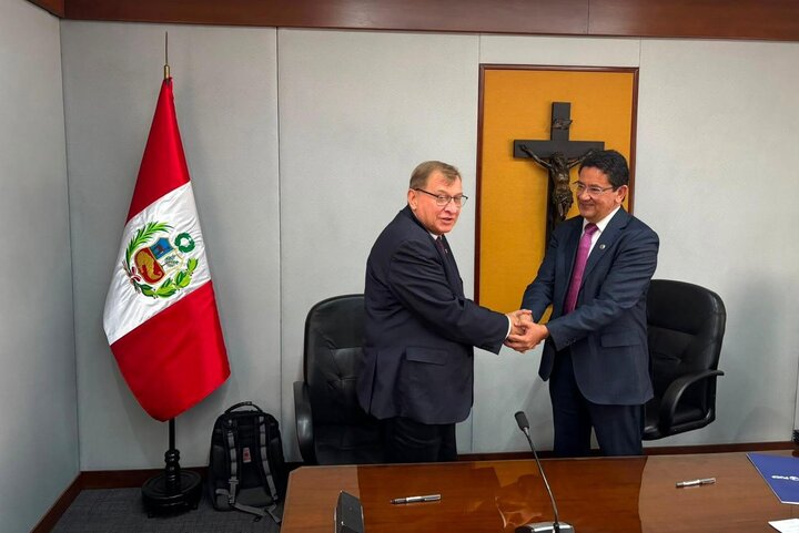 Two men in elegant attire shaking hands in a room with a large Peruvian flag and a cross.