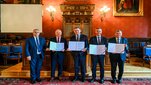 Five representatives display signed agreement documents during a formal ceremony in a historic hall.