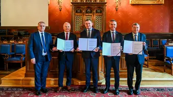 Five representatives display signed agreement documents during a formal ceremony in a historic hall.
