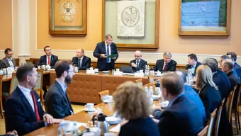 Zdjęcie przedstawia spotkanie przy dużym, prostokątnym stole w sali konferencyjnej. Uczestniczy w nim kilkanaście osób W tle widoczne są obrazy i elegancka, klasyczna aranżacja wnętrza