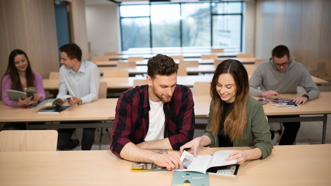 Zdjęcie przedstawia studentów i studentki, którzy siedzą przy stolikach w sali wykładowej.