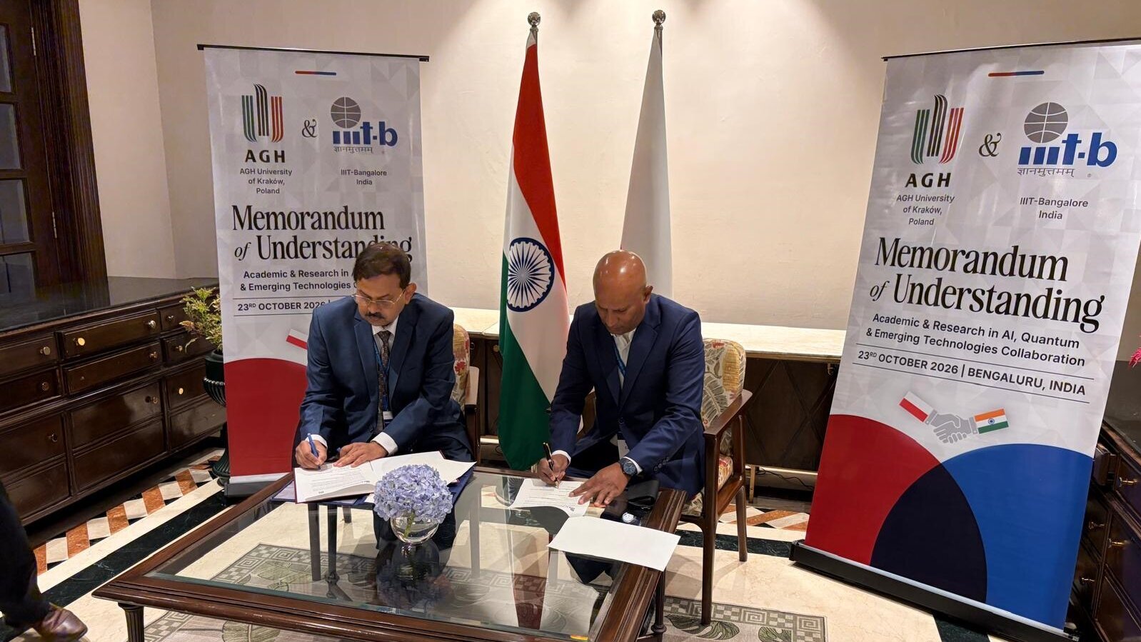 Podpisanie memorandum o współpracy między AGH a IIIT-Bangalore w Bengaluru. Przy stole siedzą sygnatariusze porozumienia, a w tle znajdują się flagi Polski i Indii oraz bannery informujące o współpracy akademickiej i badawczej.