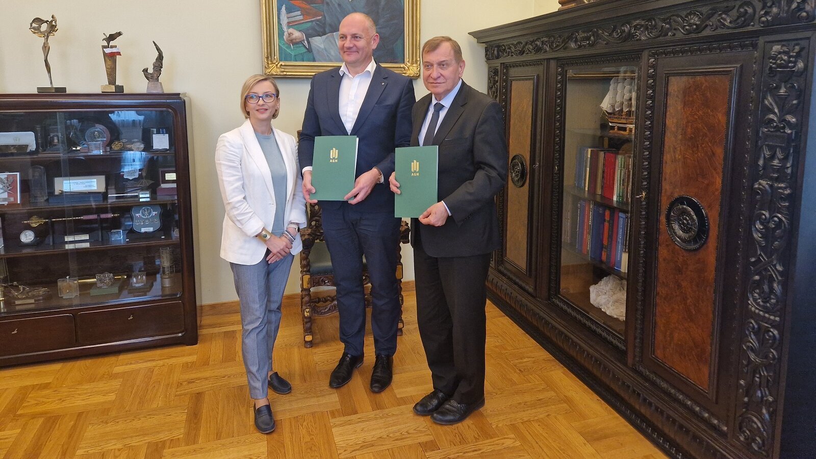 Na zdjęciu troje sygnatariuszy umowy - stoją obok siebie, Rektor AGH i prezes KPT trzymają w dłoniach zielone teczki z podpisanymi dokumentami. Zdjęcie wykonane w gabinecie rektorskim.