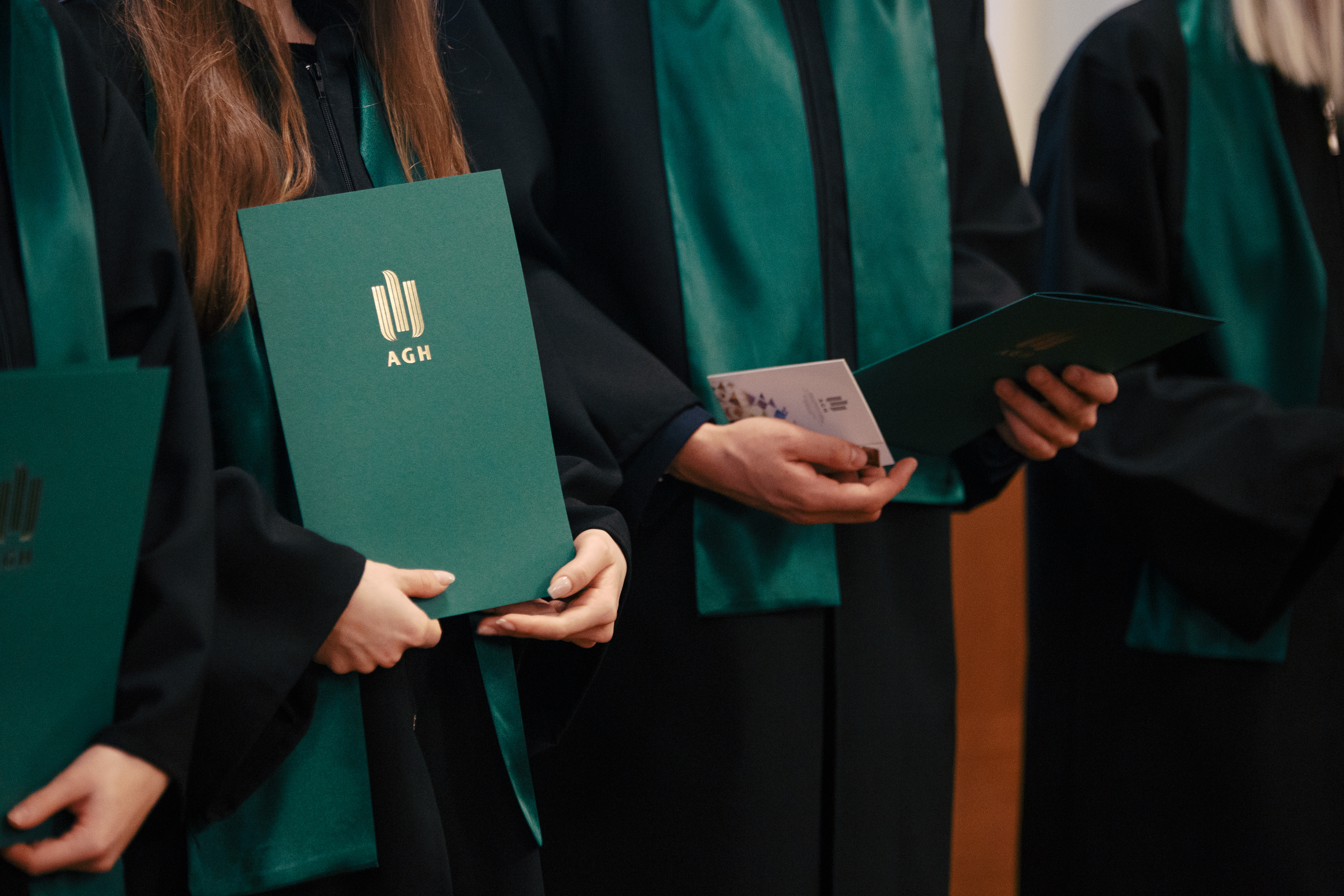 grupa studentów ubranych w togi, trzymają w rękach zielone teczki z logo AGH.