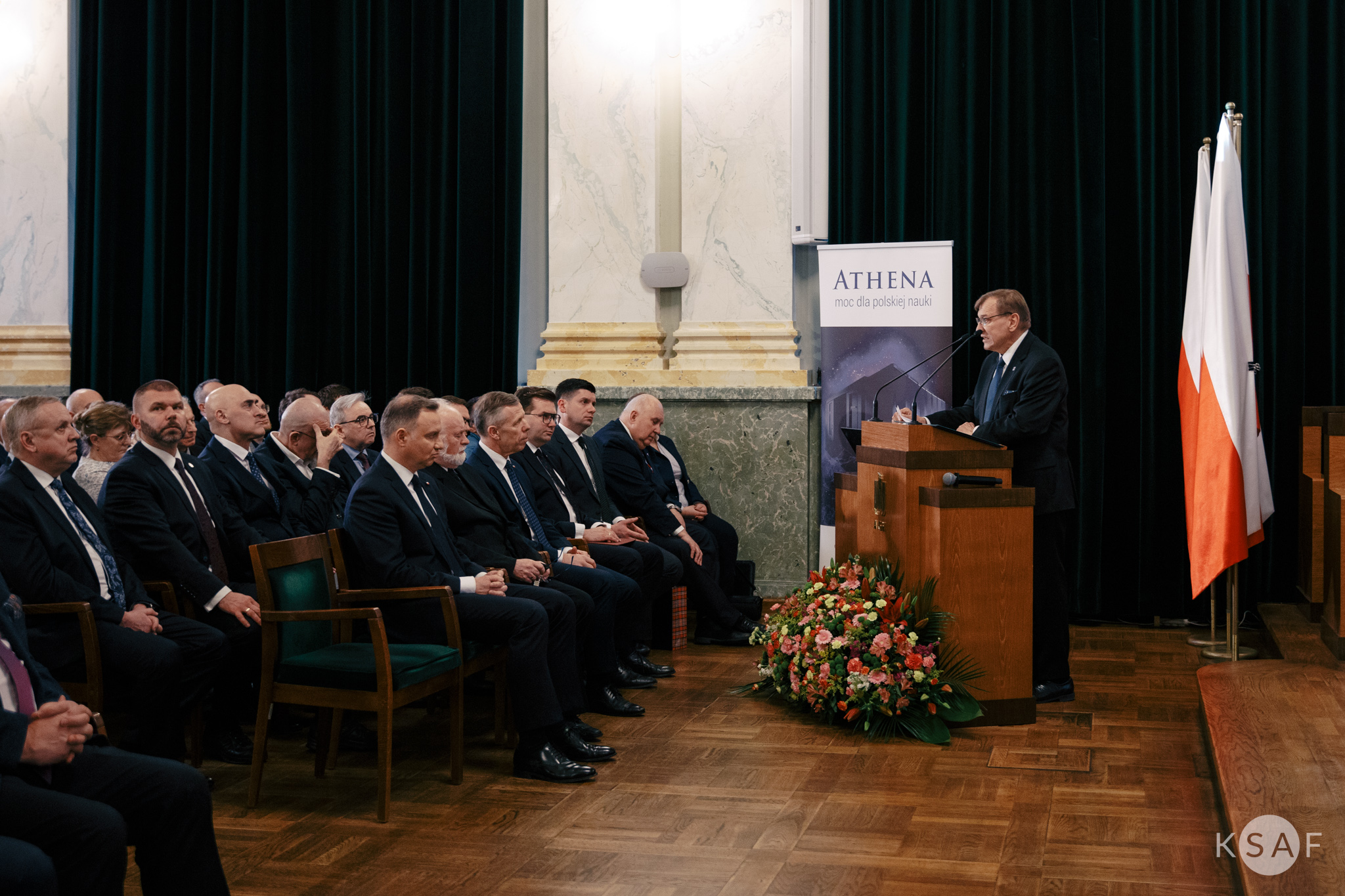 Zdjęcie przedstawia mężczyznę stojącego przy mównicy i przemawiającego do zgromadzonych w auli gości.