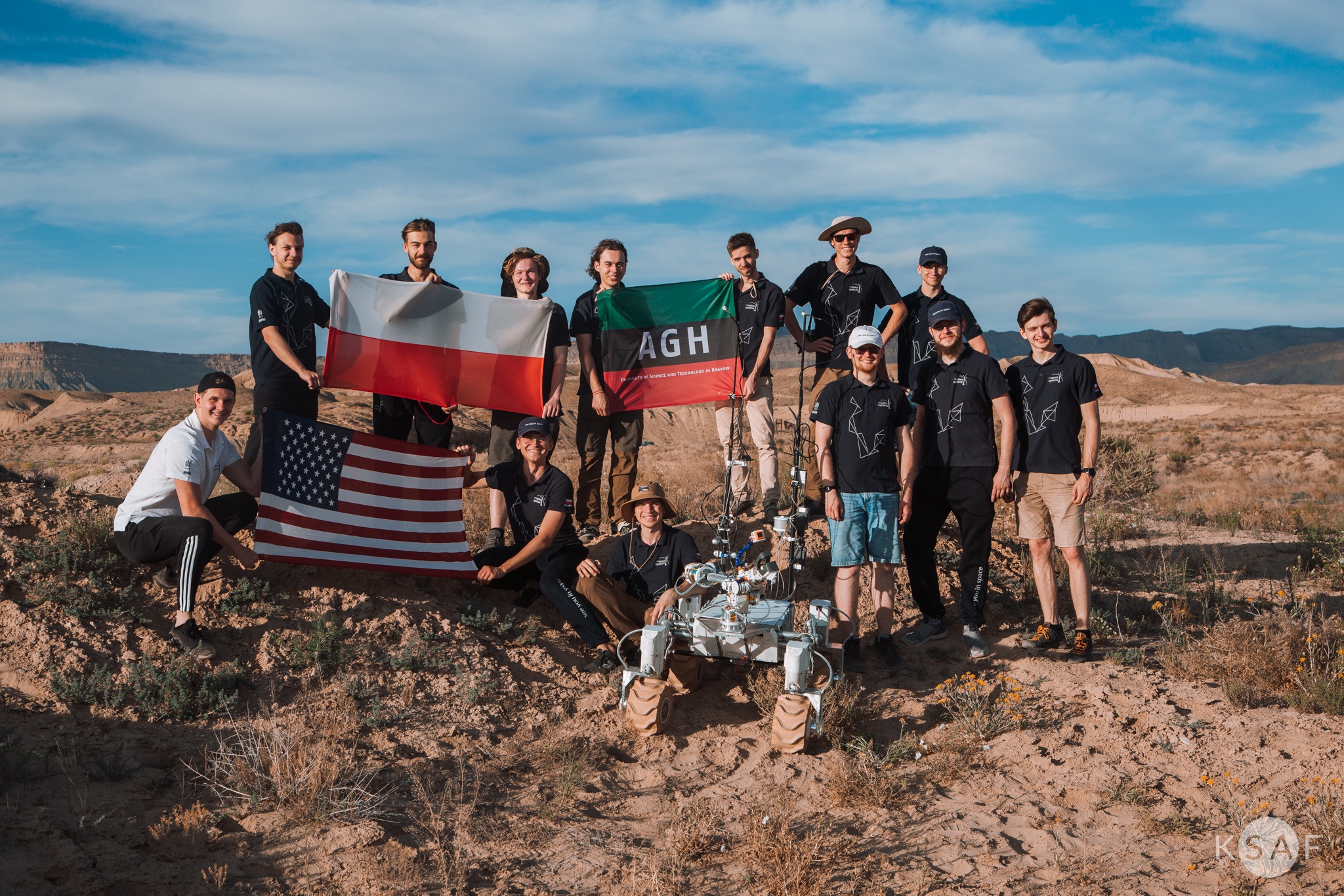 Pustynia w Utah. Grupa 11 osób - zespół AGH Space Systems, w rękach trzymają flagi Polski, USA i AGH. Przed nimi stoi łazik Kalman.
