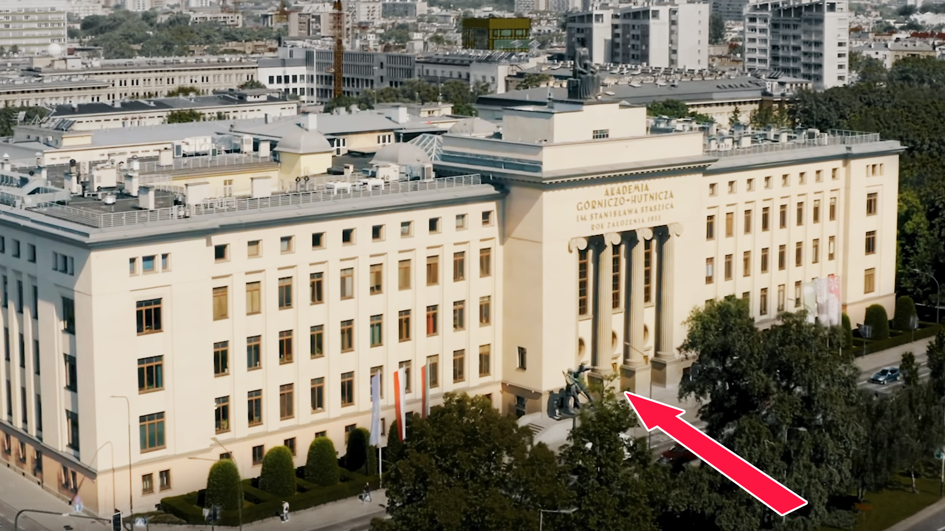 Zdjęcie z lotu ptaka przedniej elewacji budynku uczelni. Przed środkową częścią budynku umieszczono dużą czerwoną strzałkę wskazująca wejście.
