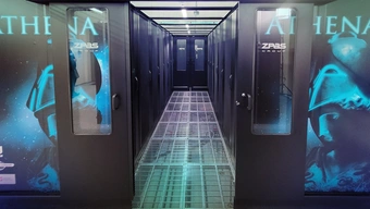 Zdjęcie korytarza utworzonego przez dwa rzędy szaf z modułami superkomputera. Korytarz po obu stronach ograniczony jest czarnymi przeszklonymi drzwiami. Podłoga korytarza jest ażurowa. Zewnętrzne boki szaf superkomputera zdobi błękitna grafika przedstawiająca głowę kobiety z założonym starożytnym hełmem. Nad sylwetką widnieje napis „ATHENA”. 