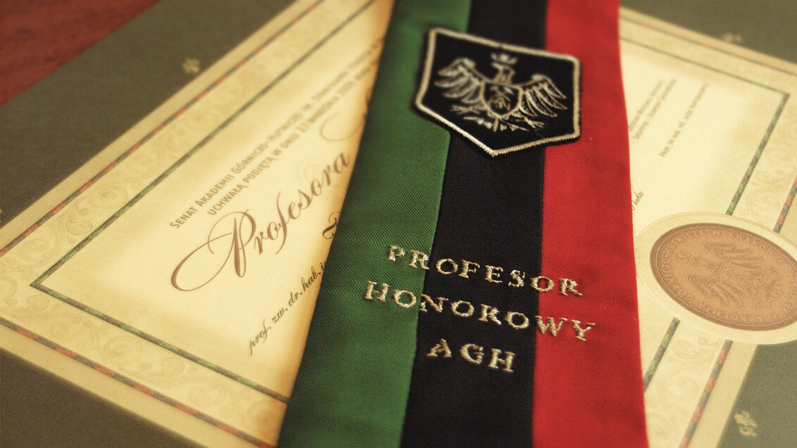 The photo shows a striped epitogium in green, black, and red with a gold inscription embroidered thereon that says: “Profesor Honorowy AGH”, which translates to AGH UST Honorary Professor. The epitogium includes the university emblem in the form of a gold eagle on a black background. Under the fabric, there is an ornamental diploma.