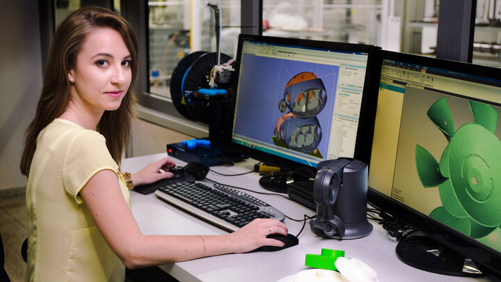 Zdjęcie zrobione w laboratorium projektowania 3d. Przy biurku, twarzą zwrócona w kierunku obiektywu siedzi młoda kobieta. W ręku trzyma mysz komputerową. Na blacie znajduje się klawiatura oraz dwa monitory. 