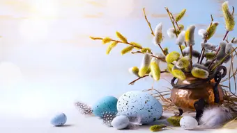 Zdjęcie kompozycji wielkanocnej. Na pierwszym planie błękitne i fioletowe jajka, za nimi metalowy wazon z kwitnącymi gałązkami wierzby.