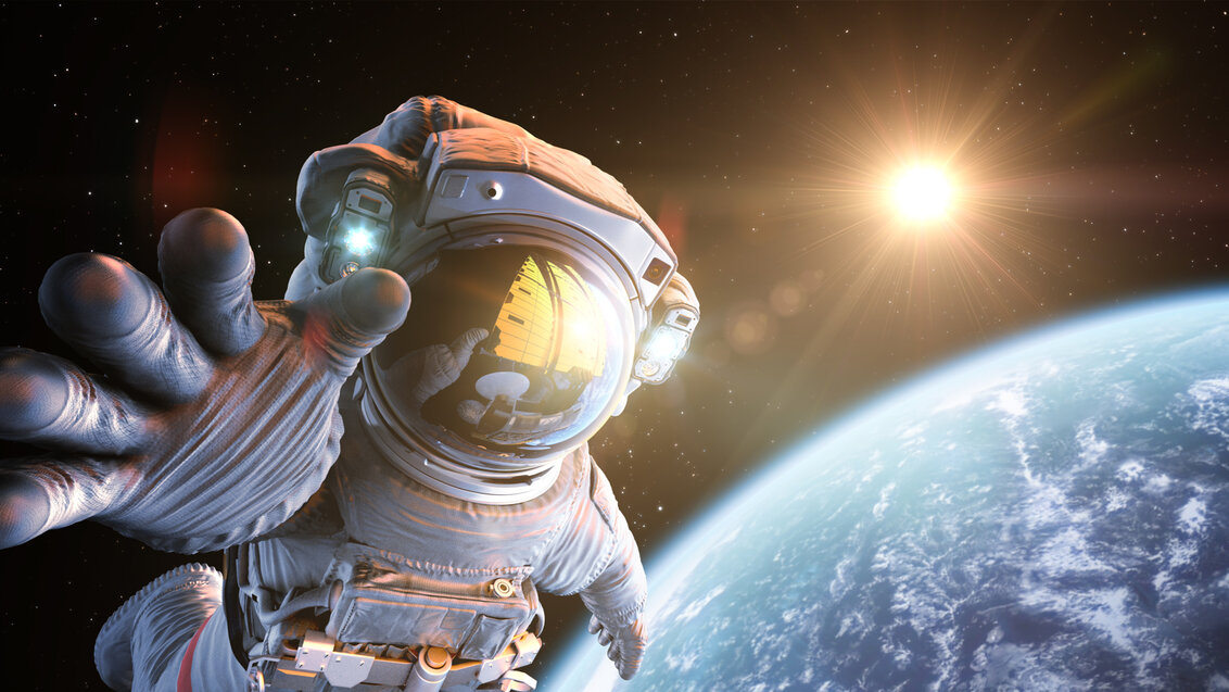 Zdjęcie przedstawiające astronautę przebywającego w przestrzeni kosmicznej, wyciągającego dłoń w stronę obserwatora. W tle kula ziemska, dalej słońce.