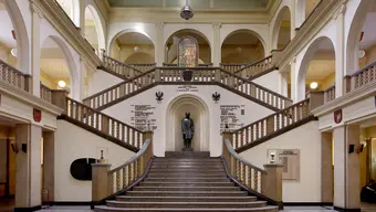 Zdjęcie wnętrza holu klasycystycznego budynku o kilku kondygnacjach. Na pierwszym planie widać centralnie usytuowane schody symetrycznie dzielące się na dwie strony. Dookoła pomieszczenia widać krużganki. Na pierwszym półpiętrze we wnęce znajduje się pomnik Staszica. Nad nim na pierwszym piętrze usytuowany jest kolorowy witraż. 