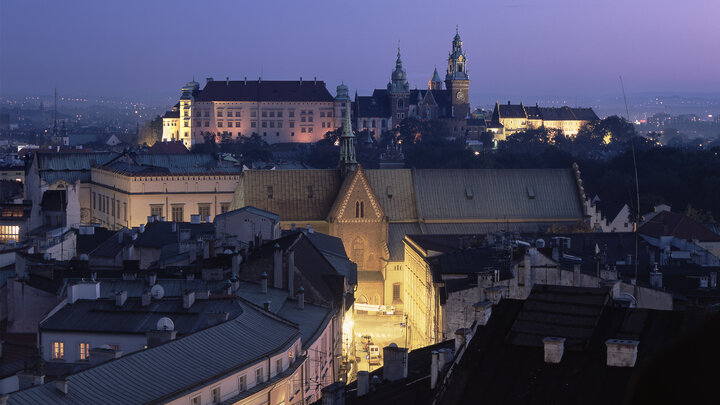 Nocne zdjęcie panoramy Krakowa utrzymane w granatowo-fioletowej tonacji. W oddali widać wzgórze z zamkiem królewskim oraz wieżami katedry. Poniżej jasno oświetlona ulica Kanonicza.
