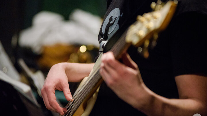 Zdjęcie przedstawia zbliżenie na fragment sylwetki gitarzysty basowego grającego na instrumencie.