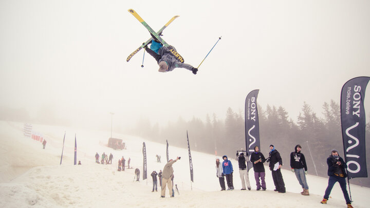 Zdjęcie przedstawia unoszącego się kilka metrów nad poziomem śniegu narciarza, wykonującego salto z prostopadle ułożonymi względem siebie nartami. Obok trasy widać publiczność.