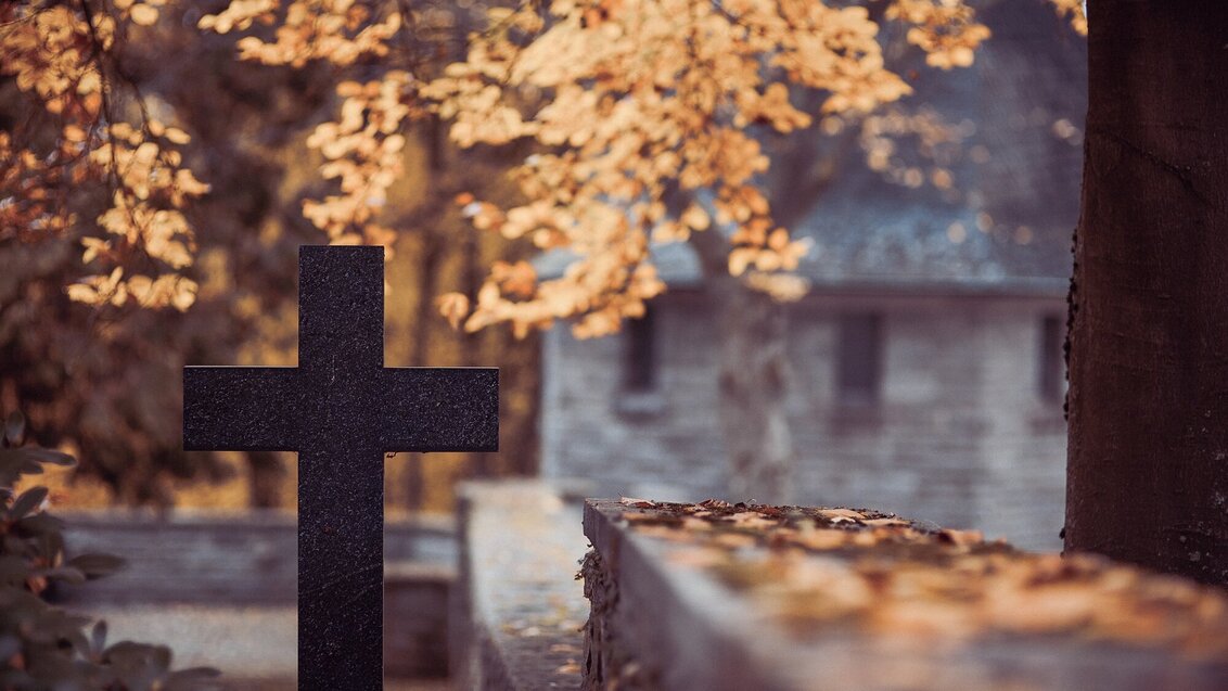 Grafika dekoracyjna. Cmentarz jesienią, widać czarny krzyż, w tle kapliczka i drzewa z żółtymi liśćmi.