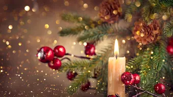 Zdjęcie przedstawia świąteczną dekorację. Na pierwszym planie płonąca świeczka, obok fragment udekorowanej choinki. W tle złote refleksy.