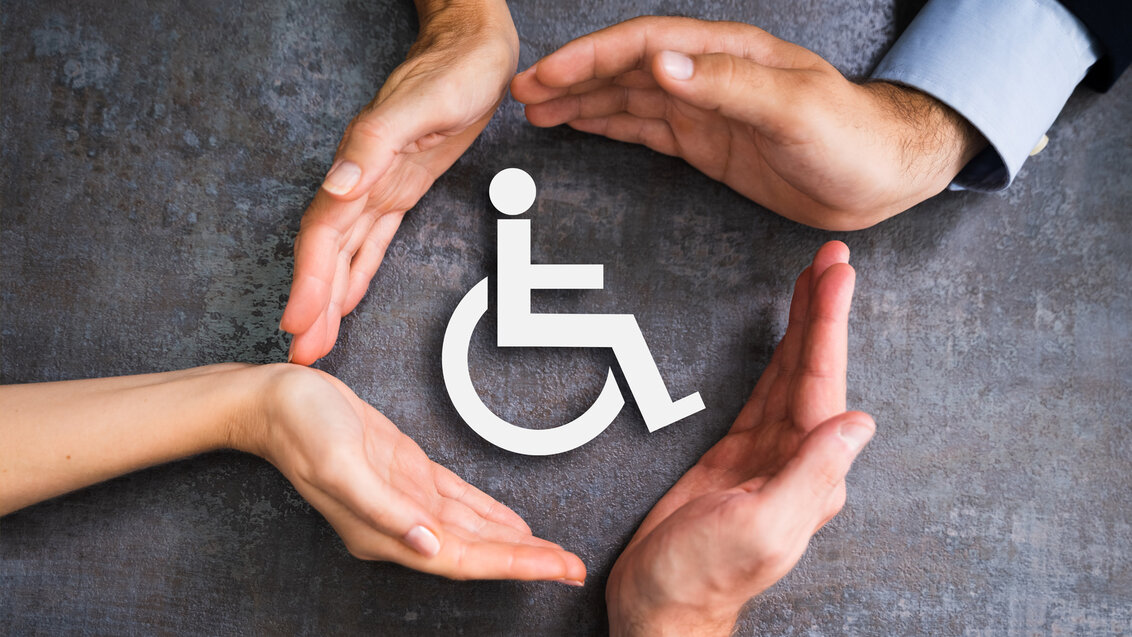 Zdjęcie przedstawiające biały znak niepełnosprawności ruchowej na ciemnym tle. Znak otoczony jest przez cztery dłonie.