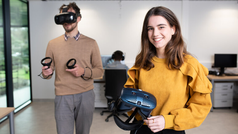 A photo of two young people in a spacious room. The man on the left is wearing VH goggles and using two manipulators. The woman on the right holds VR goggles and looks at the observer.