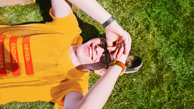 Zdjęcie młodego mężczyzny leżącego na plecach na trawie w słoneczny dzień. Mężczyzna uśmiecha się i rękami zasłania oczy przed oślepiającym go światłem. Nosi żółty t-shirt. Pod głową znajdują się jego buty.