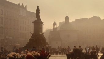 The photo shows Krakow's main square in low air transparency conditions. In the foreground, there is a statue of a man. In the background, there are outlines of historic tenements. There are passers-by on the square.