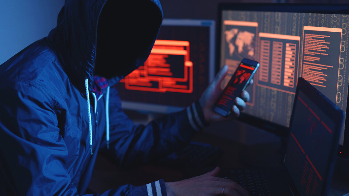 Zdjęcie przedstawia górną część sylwetki zakapturzonego mężczyznę. Mężczycnę otaczają ekrany komputerów. W dłoni trzyma smartfon. Ekrany wyświetlaja dane.