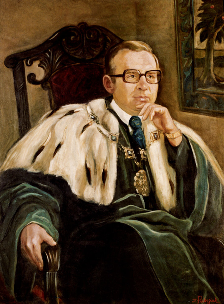 Portret przedstawiający Rektora siedzącego w fotelu. Rektor ma na sobie togę w zielonym kolorze, duży gronostajowy kołnierz oraz ozdobny łańcuch. Nosi okulary.