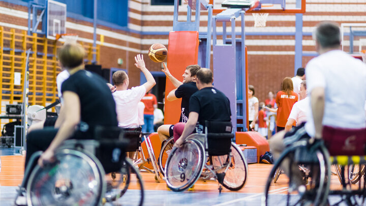 Zdjęcie niepełnosprawnych koszykarzy poruszających się na wózkach. Zawodnicy jednej z drużyn ubrani są w czarne stroje, drugiej z jasne. Jeden zawodnik próbuje przejąć piłkę w trakcie podania drugiego.