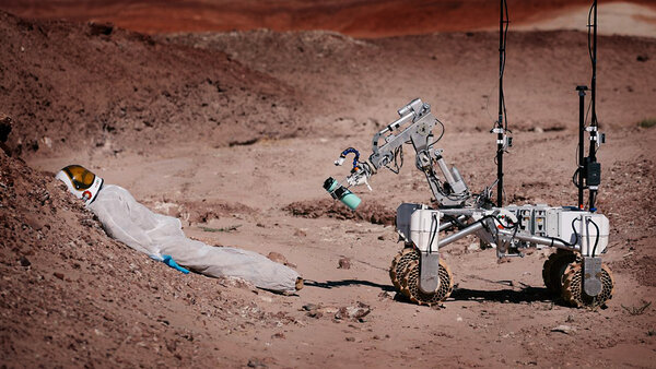 Zdjęcie przedstawia krajobraz marsjański w którym łazik marsjański zbliża się w stronę leżącego nieruchomo astronauty. W mechanicznym ramieniu pojazdu widać niebieski bidon.