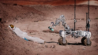 The photo shows a Martian landscape and a rover approaching a motionless astronaut dummy. The mechanical arm of the robot holds a blue water bottle.