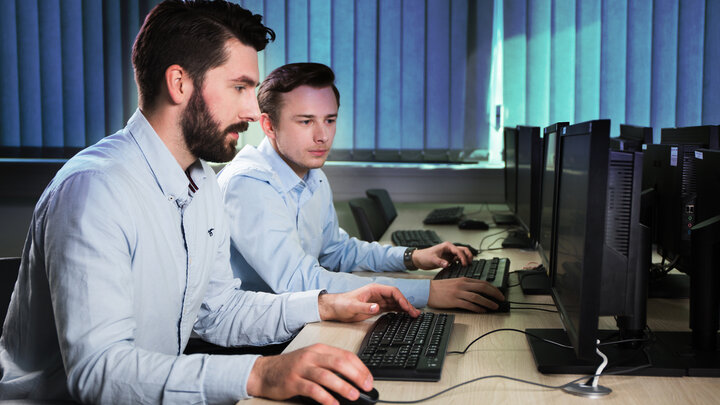 Zdjęcie przedstawia dwóch mężczyzn siedzących przy podłużnym biurku. Pracują przy komputerach i spoglądają w monitory. Siedzący bliżej posiada brodę i wąsy. Oddalony od obserwatora jest ogolony. Obaj mają na sobie koszule w błękitnym kolorze. W tle widać pionowe niebieskie rolety zasłaniające okna.