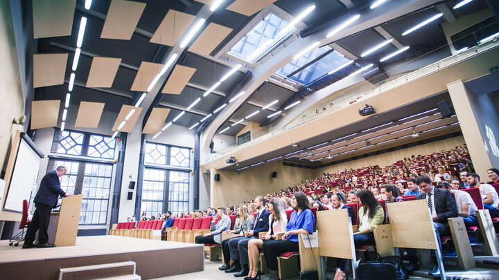 Zdjęcie dużej sali audytoryjnej w któej odbywa się wykład. Po lewej stornie widać prelegenta stojącego na katedrze. Po prawej stronie w rzędach siedza studenci. 
