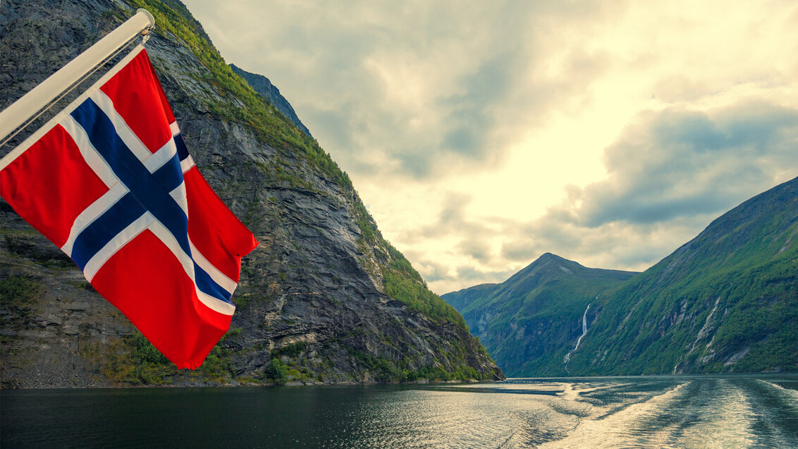 Zdjęcie przedstawia zbliżenie na norweską flagę (granatowy krzyż z białym obramowaniem na czerwonym tle) na tle panoramy fiordów. W dole widoczny ślad torowy statku, po bokach strome brzegi zatoki.