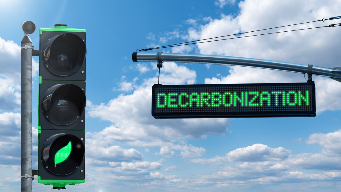Abstrakcyjne zdjęcie przedstawiające z lewej strony drogowy sygnalizator świetlny z podświetlonym światłem zielonym w kształcie liścia. Po prawej widoczny wiszący wyświetlacz LED ze słowem „DECARBONIZATION”.