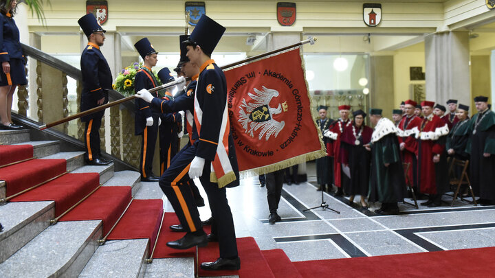 Two students in ceremonial uniforms carry a red banner with a white eagle in a crown in the centre. In the background, an assembly of deans in ceremonial attire.