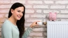 Zdjęcie przedstawia młodą kobietę trzymająca dłoń na termostacie grzejnika ściannego. Na grzejniku postawiona jest różowa świnka skarbonka.