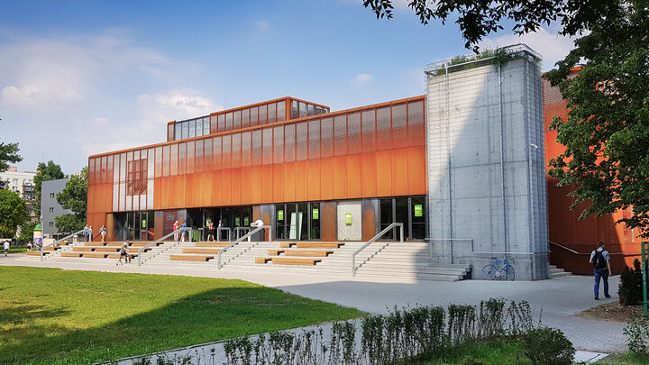 Zdjęcie nowoczesnego budynku o surowej elewacji wykończonej pomarańczową skorodowaną blachą kortenową z która kontrastują betonowe elementy ściany oraz podłużne schody prowadzące do wejść budynku. Na schodach usytuowano rzędy drewnianych ławek. Przed budynkiem znajduje się spory zieleniec. 