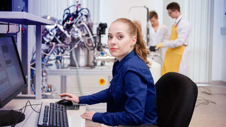 Zdjęcie przedstawia trzyosobową grupę naukowców w laboratorium inżynierii materiałowej. Na pierwszym planie widać siedzącą przy biurku młodą kobietę. Trzyma w ręku mysz komputerową. Na blacie znajduje się klawiatura oraz monitor. Kobieta siedzi bokiem do obserwatora i spogląda w jego stronę. W tle po lewej znajduje się skomplikowana aparatura badawcza zbudowana z błyszczących metalowych elementów rozgałęziających się we wszystkich kierunkach w pajęczynie różnokolorowych przewodów. W tle po prawej dwóch młodych mężczyzn pracujących przy sprzęcie.