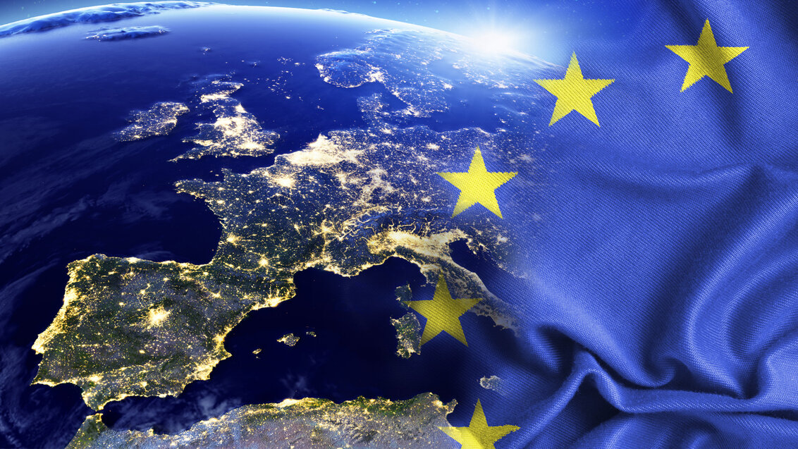 Abstrakcyjne zdjęcie przedstawiające widok fragmentu Europy widziany nocą z przestrzeni kosmicznej. Po prawej stronie widać zarys flagi Unii Europejskiej oraz blask wschodzącego słońca.