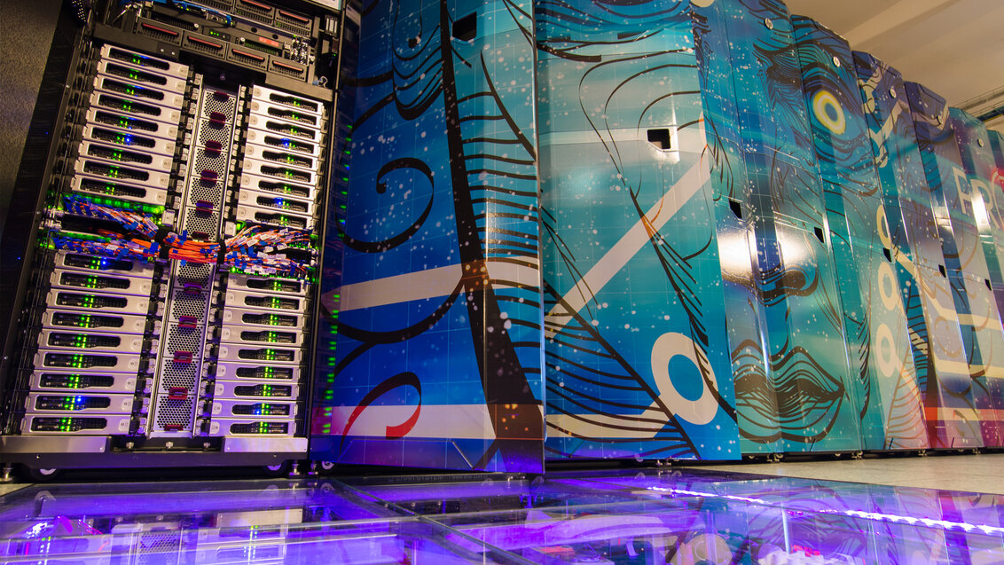 Zdjęcie przedstawia rząd pionowych szaf w których znajdują się podzespoły superkomputera. Drzwi szaf ozdobione są abstrakcyjną grafiką przedstawiającą ludzką twarz ze świecącymi się oczyma. W pierwszej, otwartej szafie widać kolumnę jednakowych modułów. Na środku wysokości widoczne kolorowe wiązki przewodów. Fragment podłogi jest przezroczysty i podświetlony fioletowym światłem. 